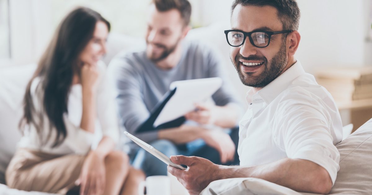 Confident sales manager. Cheerful mature man holding digital tablet and looking over shoulder while another man and woman sitting in the background