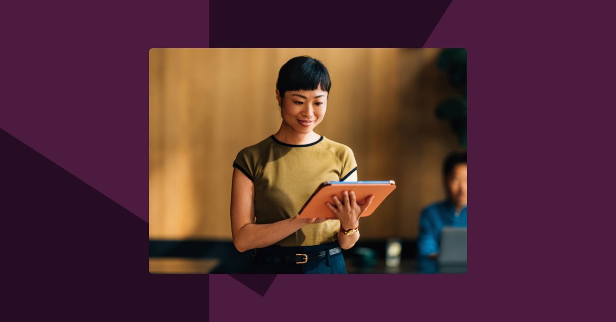Woman standing looking at ipad in an office with a colleague out of focus seated behind her