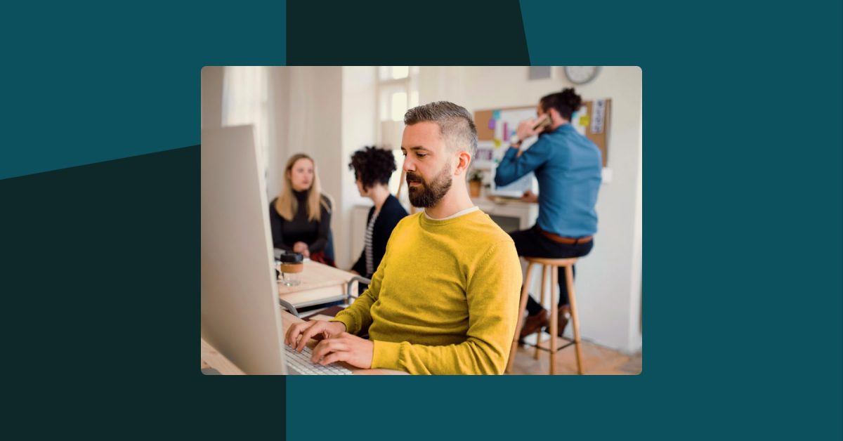Man working on a computer monitor in an office with out of focus colleagues behind him