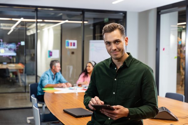 Portrait of confident young caucasian businessman with tablet pc while colleagues in background. Unaltered, creative business, workplace, meeting, wireless technology.