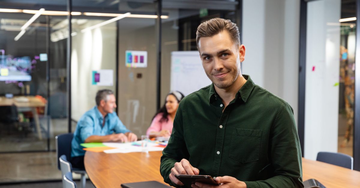 Portrait of confident young caucasian businessman with tablet pc while colleagues in background. Unaltered, creative business, workplace, meeting, wireless technology.