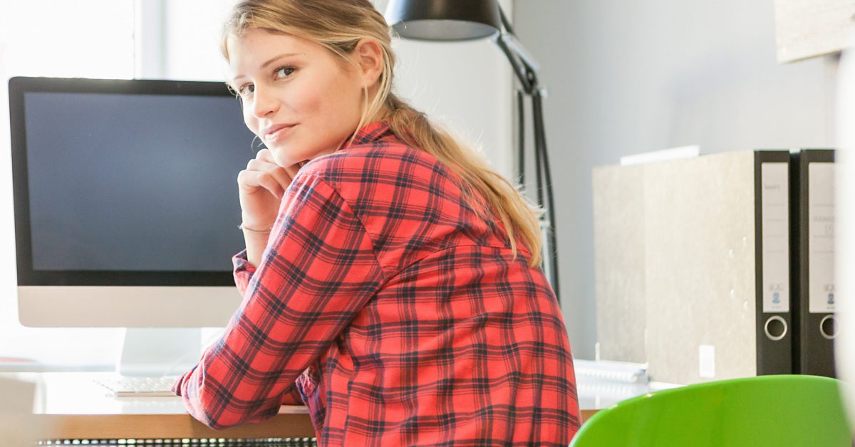 Rear view of young woman at computer looking over shoulder at camera smiling
