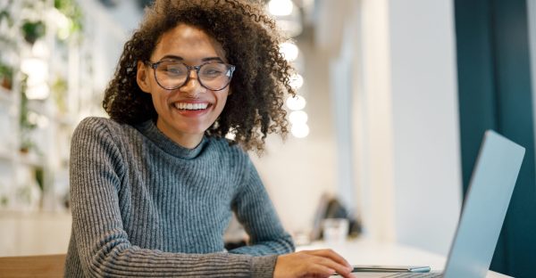 Smiling african woman freelancer working laptop while sitting in coworking and looking at camera