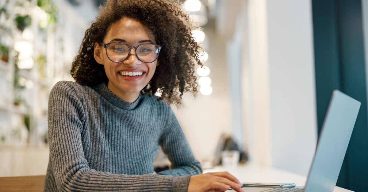 Smiling african woman freelancer working laptop while sitting in coworking and looking at camera