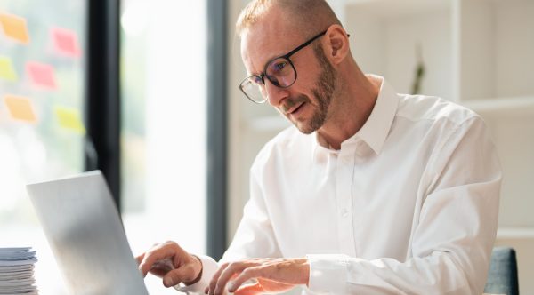 Young business man wearing glasses using laptop, typing on keyboard, writing email or message, sitting in office.