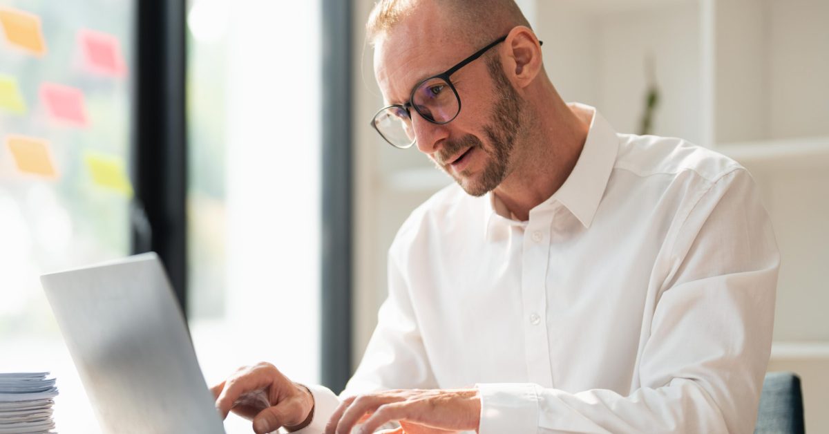 Young business man wearing glasses using laptop, typing on keyboard, writing email or message, sitting in office.