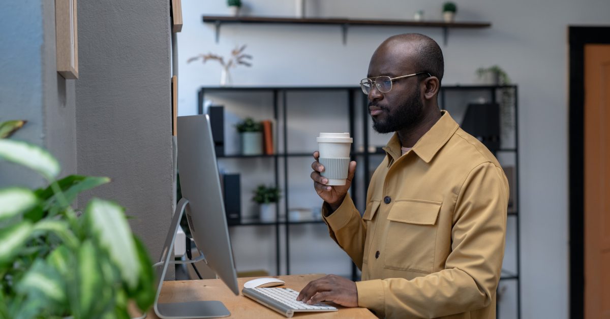 Young serious employee having drink and looking at data on computer screen while standing by workplace in urban office